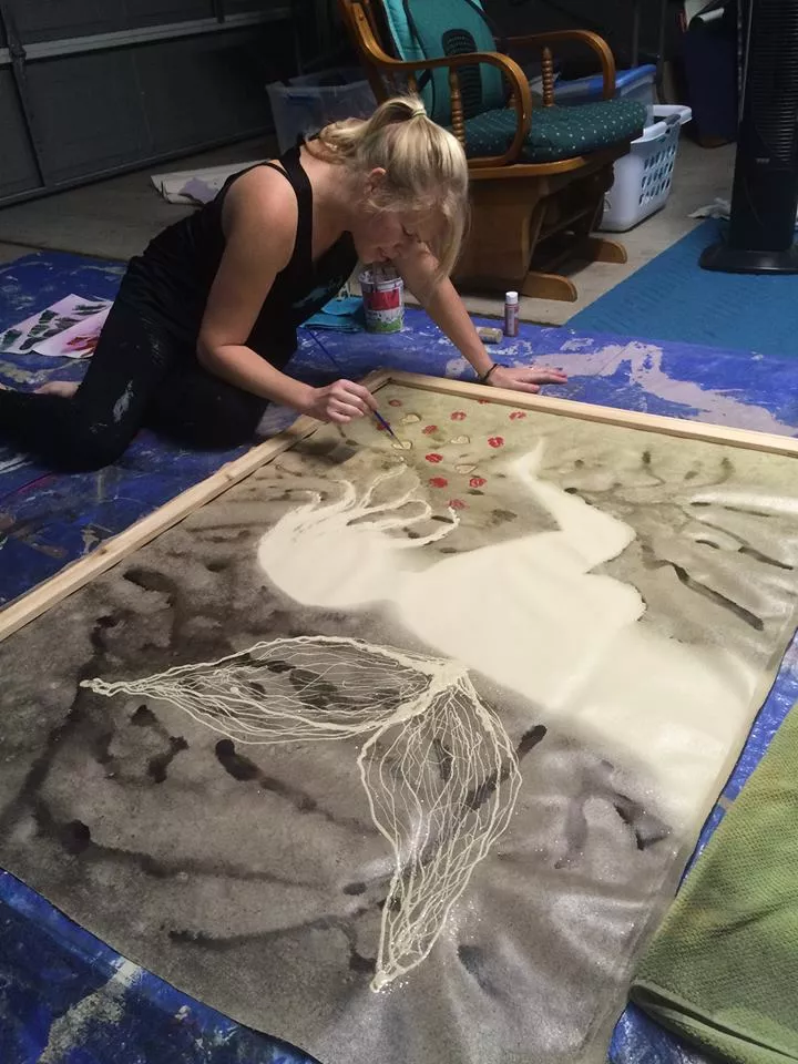 Person painting an abstract design on a large canvas on the floor, with various art supplies around.