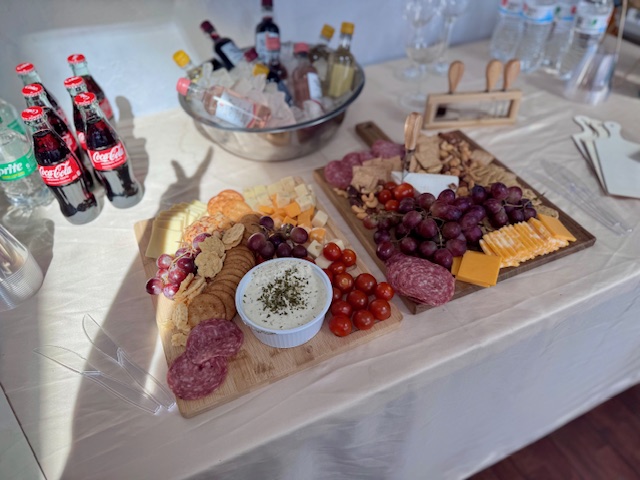 Charcuterie boards with salami, cheese, grapes, cherry tomatoes, and crackers adorn the table at Bunnie's Business. Nearby are assorted drinks, including bottles of soda and water, setting the stage for a delightful painting event.