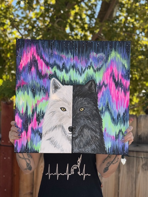 A person holds a striking painting of a wolf, its face split into white and black to symbolize duality, set against a vibrant aurora borealis. Their own face is hidden behind the artwork—perfect inspiration for your next Painting Event.