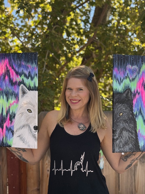 A woman stands outdoors holding two paintings of wolf heads, one white and one black, against colorful abstract backgrounds—symbolizing duality. She is smiling and wearing a black tank top with a graphic design at a fundraising painting event.