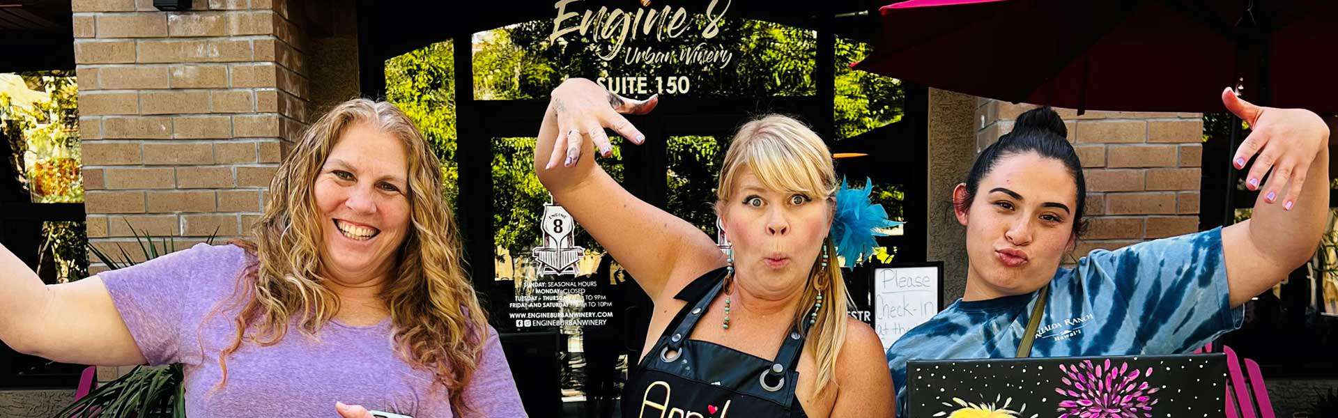 Three women, standing outside a storefront, making playful gestures towards the camera. The middle woman wears an apron with "Amy" on it, while the other two wear casual clothing.