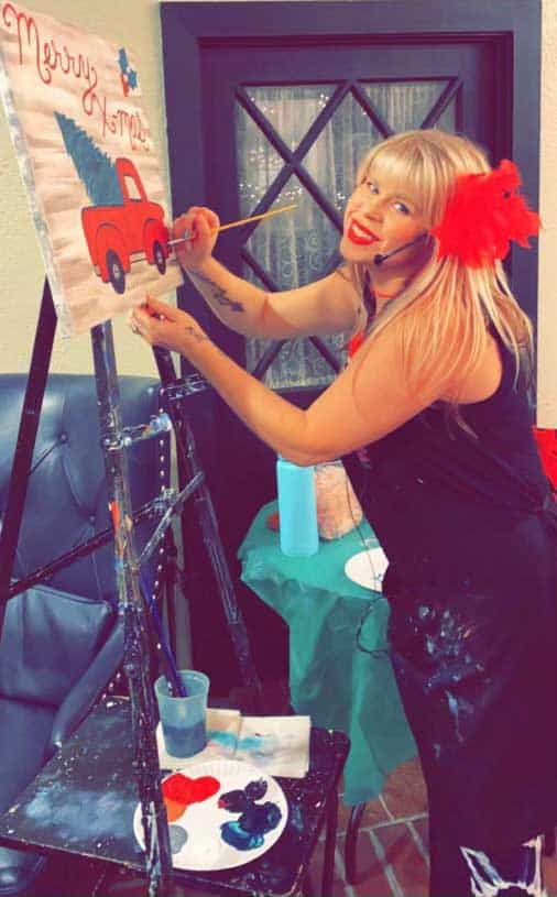 A person with long hair and a red flower in their hair is painting a picture of a red truck and Christmas tree on a canvas. They are wearing a black outfit and standing next to a paint palette and easel.