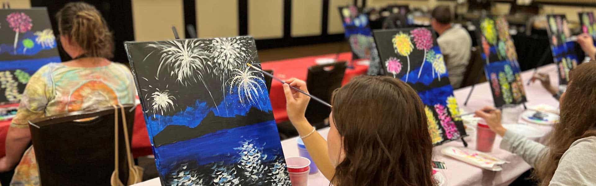 People sitting at tables painting fireworks on canvases in an indoor art class setting.