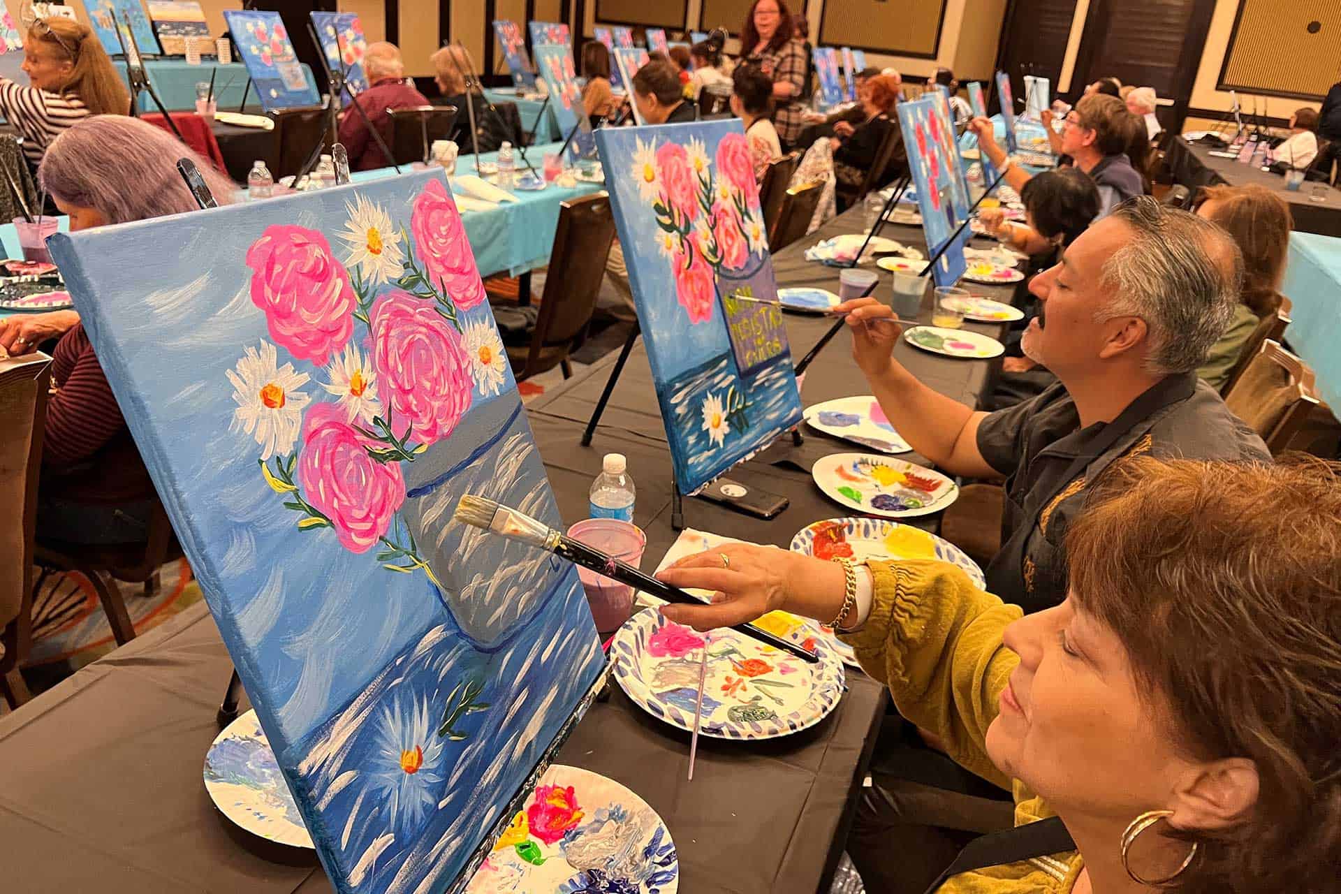 A group of people sit at tables in a room, painting similar floral artworks on canvases. Each person has painting supplies and drinks on the table in front of them.