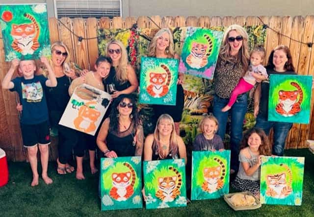 A group of people pose outdoors, each holding a painting of a cartoon tiger. The group includes children and adults, with colorful paintings and a wooden fence in the background, showcasing their artistic achievements from Service 2.