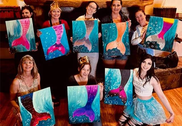 A group of nine women displays their colorful paintings of mermaid tails while standing together in a room. Some wear crowns, and they pose cheerfully with their artwork, celebrating their shared creative service to the community.