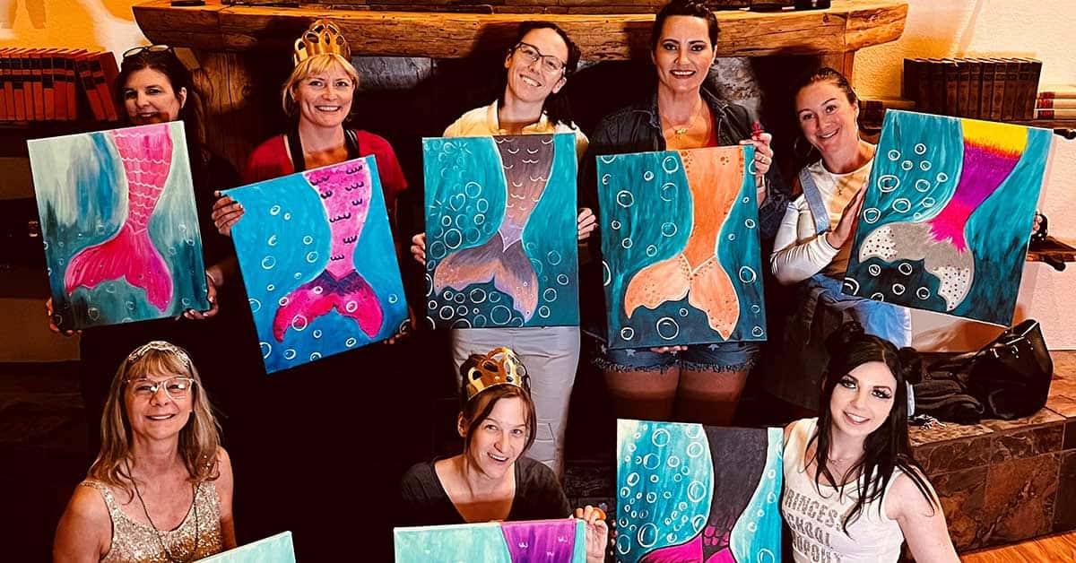 A group of women indoors, some wearing crowns, joyfully engage in private painting events. They proudly display their artwork featuring mermaid tails and bubbles.