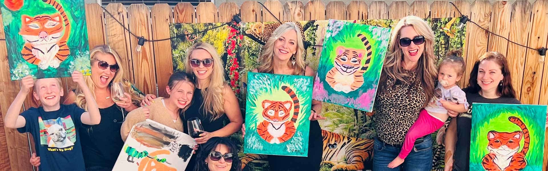 A group of people holding paintings of tigers stand in front of a wooden fence outdoors. Some hold glasses and a child is present. One person's dog is seen at the bottom.