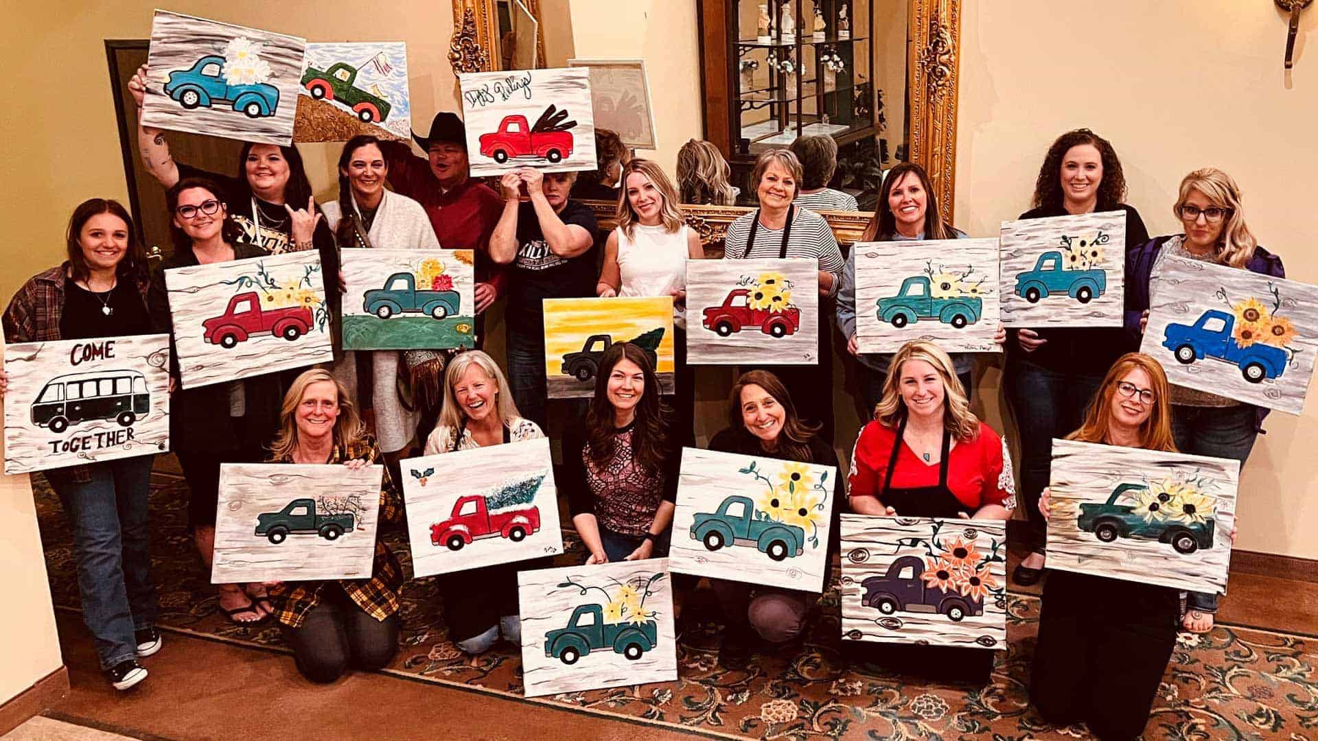 A group of people posing indoors, each holding a painting of a car. The paintings feature different designs and colors. A large mirror and ornate frame are visible in the background.