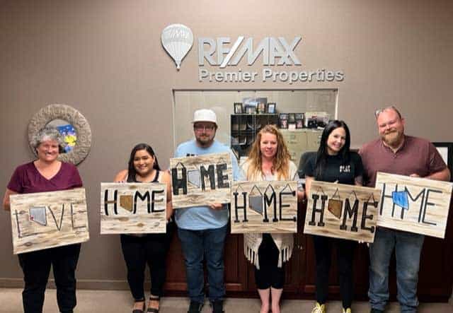 Six individuals standing in a row hold up wooden signs with "LOVE" and "HOME" in front of a RE/MAX Premier Properties office logo on the wall, showcasing their commitment to exceptional service.