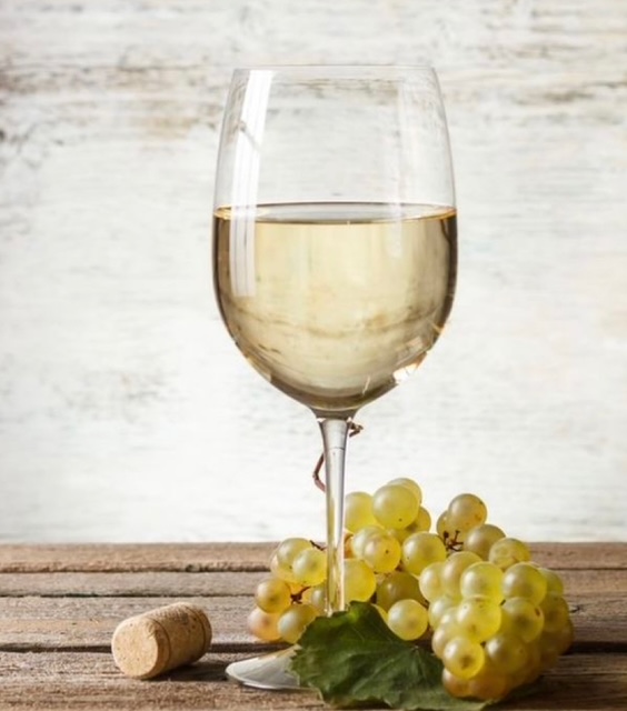 A glass of white wine rests on a wooden surface, complemented by a bunch of green grapes and a wine cork, reminiscent of a still life captured at an artful painting event.